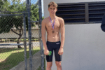 Young male swimmer wearing black racing jammers stands outdoors with a medal around his neck beside a chain-link fence.