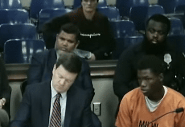 Courtroom image shows a man in an orange jail uniform seated beside an attorney, with officers and spectators behind them.