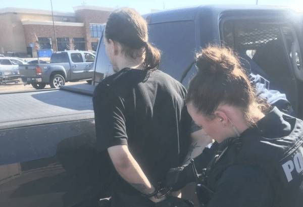 A person stands beside a vehicle while a uniformed police officer appears to conduct an interaction in a parking lot.