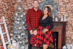 Family of three poses in front of a decorated Christmas tree and fireplace, wearing coordinated red-and-black holiday outfits.