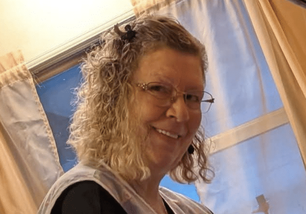 Smiling woman with curly blond hair and glasses poses indoors near a window with light-colored curtains.